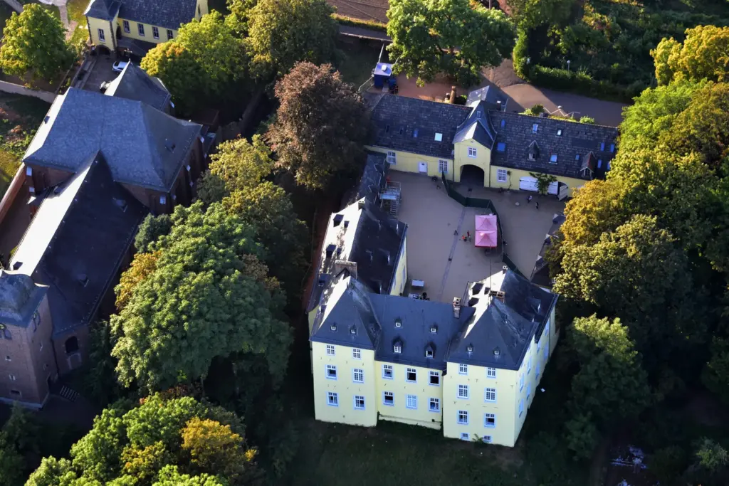 Luftbild von Schloss Alfter mit gelber Fassade und Innenhof zwischen alten Bäumen