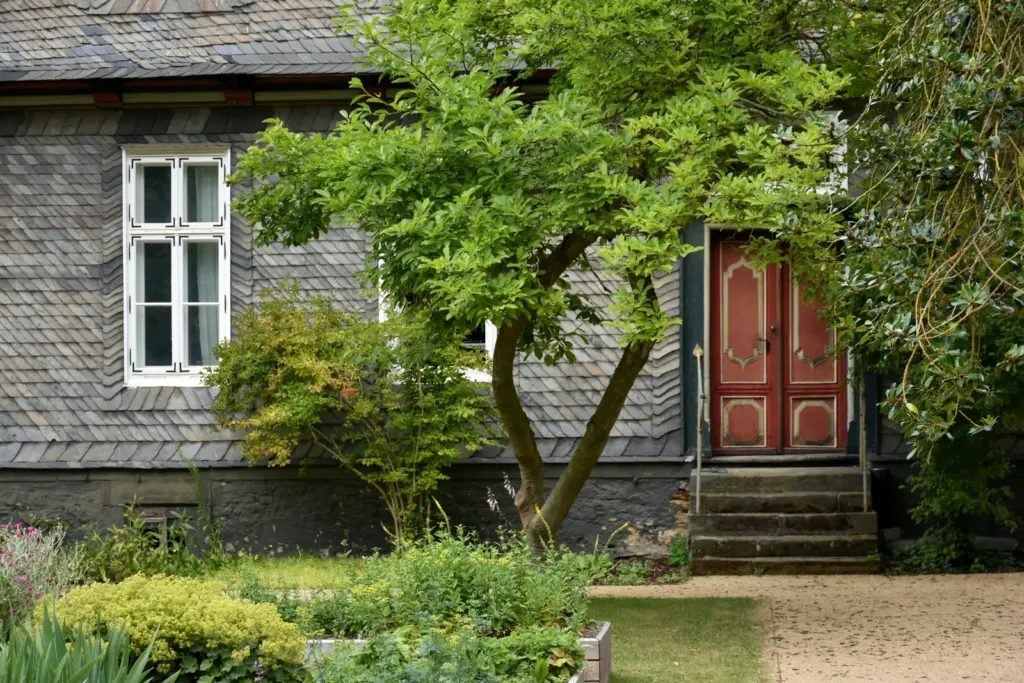 Älteres Wohnhaus mit Schieferfassade, roter Haustür und grünem Garten mit Baum